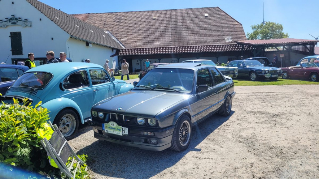 Eine Ansammlung klassischer Autos auf einem Freigelände. Ein blauer VW Käfer und ein grauer BMW E30 sind nebeneinander geparkt. Im Hintergrund tummeln sich Menschen um die Autos herum.